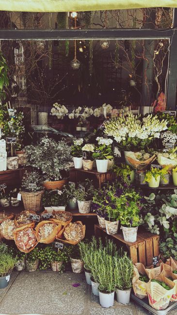 A display of fresh flowers at Flowers Acton, featuring a variety of blooms including white lilies, white roses, and purple irises arranged in bouquets and potted plants. The flowers are arranged on wo