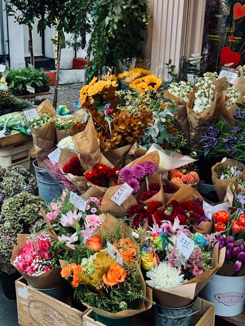 A vibrant outdoor display of fresh flower bouquets arranged on tables and in buckets, featuring a variety of blooms such as bright yellow sunflowers, pink lilies, red roses, purple irises, and white c