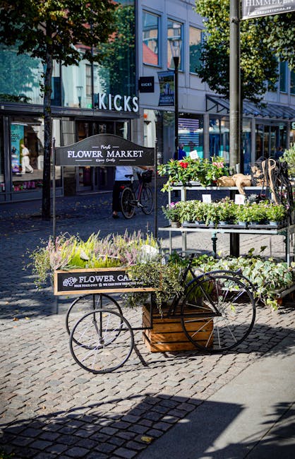An outdoor flower cart on a cobblestone sidewalk displays fresh flowers in various colors and varieties, including lavender, white, and green foliage, arranged in black metal trays and wooden boxes. T