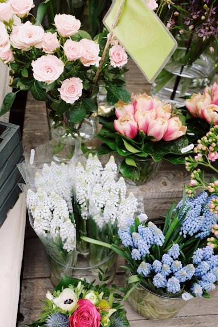 A display of fresh floral arrangements placed on a rustic wooden surface, featuring various types of flowers including light pink roses in a tall clear glass vase, pale pink tulips with some yellow-ti