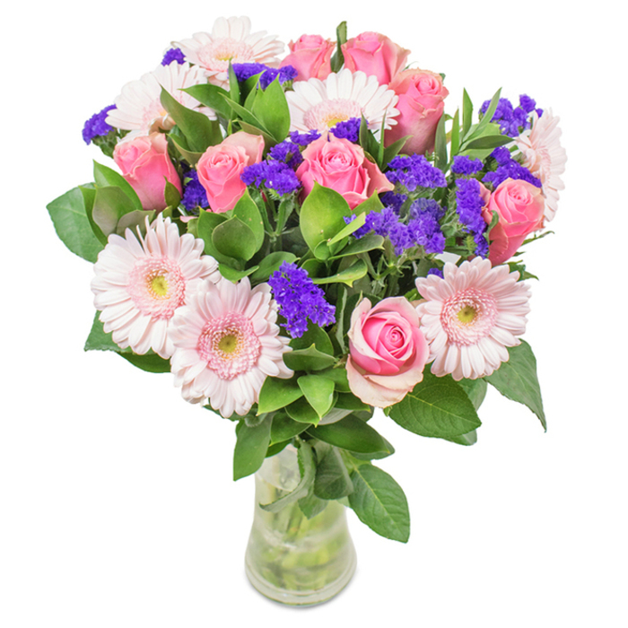 An exquisite bouquet arranged by a skilled Acton florist, displayed in a tall, slender glass vase with visible stems and clear flowing water, set against a neutral white backdrop. The centre of the bouquet draws the eye: approximately eight elegantly formed pink roses, their petals just beginning to open in classic spiral formation. These harmonise gorgeously with light pink Gerbera daisies, five or six strong, whose-wide faces and almost-white centres add both softness and volume. Sprigs of lively purple statice-known for their distinctive papery texture and bold hue-are interspersed for a striking pop of colour, referencing the vibrant floral stalls at Acton Market. A base of abundant deep green foliage provides not only fullness, but also an inviting, earthy foundation. The bouquet embodies freshness, refinement, and gentle celebration, making it an ideal choice for marking anniversaries, birthdays, or simply to enliven a sitting room along Churchfield Road. It resonates with the joy and appreciation for beauty that defines the Acton community.